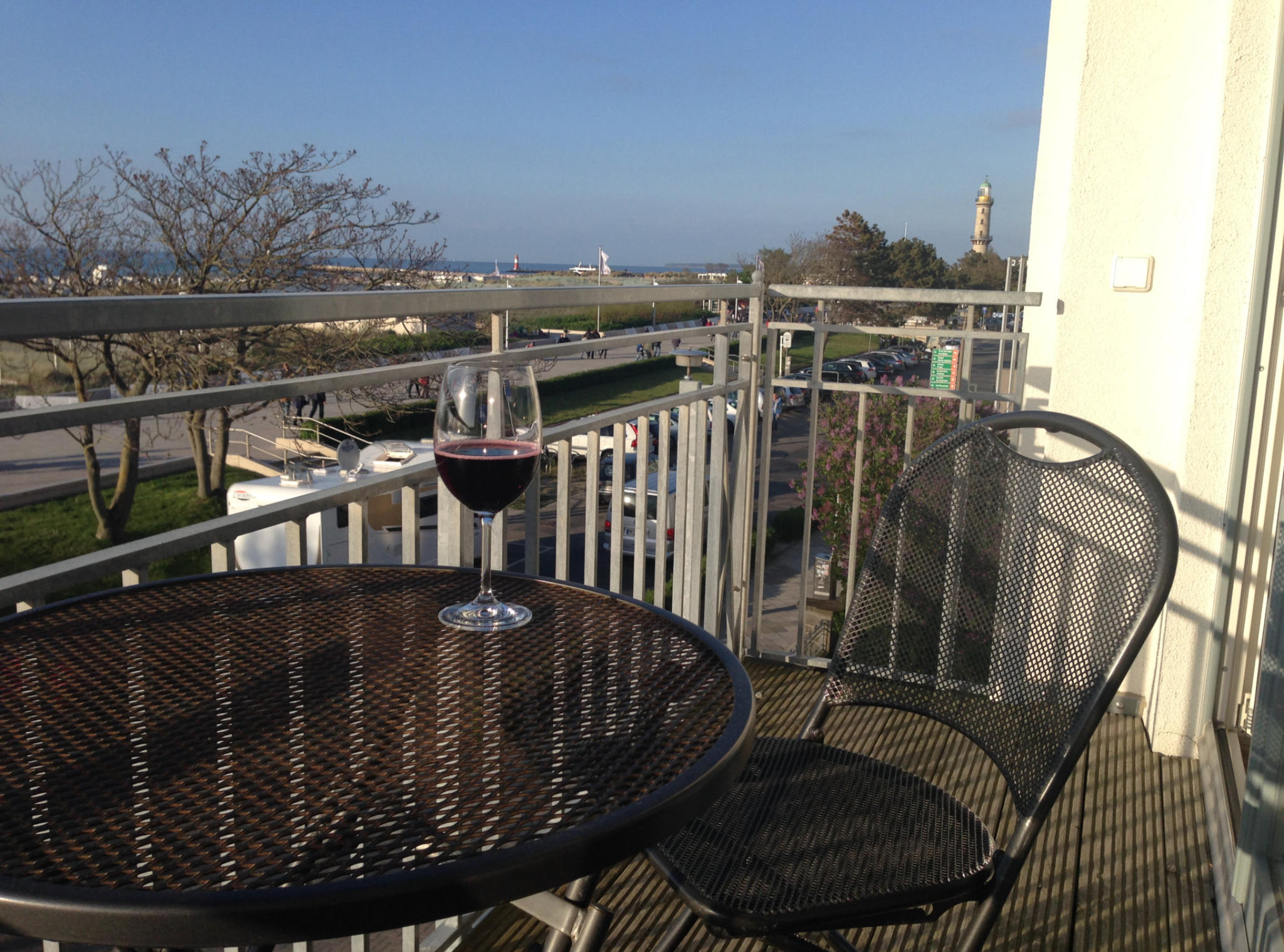 Balkontisch mit Weinglas, dahinter Seepromenade, Ostsee und der Leuchtturm von Warnemünde